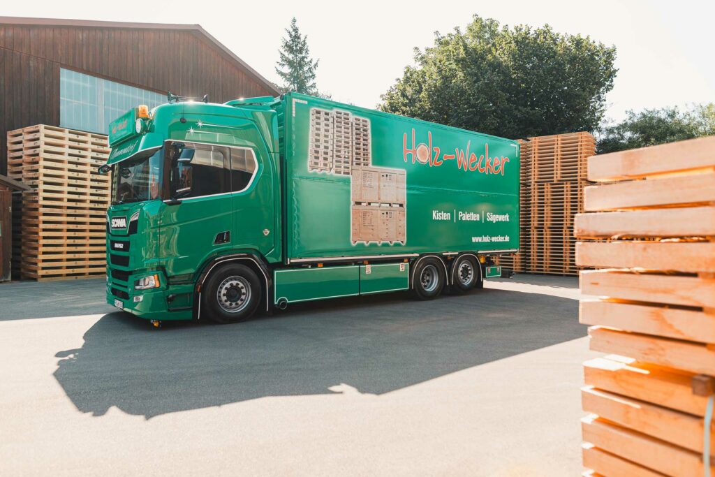 Holz Wecker LKW in Grün auf dem Firmengelände. Im Hintergrund Paletten und Schnittholz.